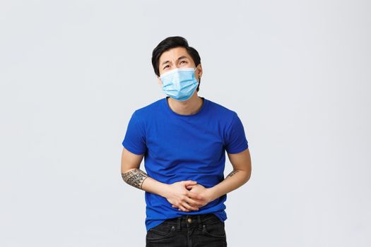 Different emotions, social distancing, self-quarantine on coronavirus and lifestyle concept. Asian man in medical mask feeling sick, have stomachache, catch food poison or flu.