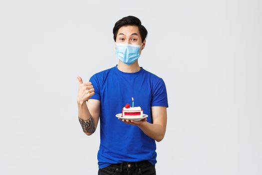 Self-quarantine, home lifestyle and celebration concept. Happy satisfied asian man ordered home delivery of birthday cake, thumb-up as holding it, wear medical mask, pleased with takeaway.