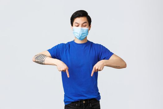 Different emotions, social distancing, self-quarantine on coronavirus and lifestyle concept. Excited and intrigued asian man in medical mask, pointing fingers down and looking at banner curious.