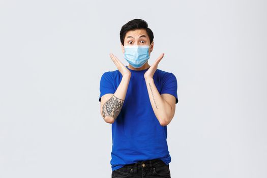 Different emotions, social distancing, self-quarantine on coronavirus concept. Surprised and astonished, excited asian man in blue t-shirt and medical mask, gasping and staring impressed.