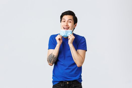 Different emotions, social distancing, self-quarantine on covid-19 and lifestyle concept. Cheerful handsome asian guy take-off medical mask to smile you, standing grey background.