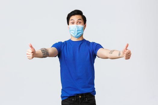 Different emotions, social distancing, self-quarantine on covid-19 and lifestyle concept. Surprised cheerful asian man in medical mask and t-shirt, reaching for hug, greeting guest.