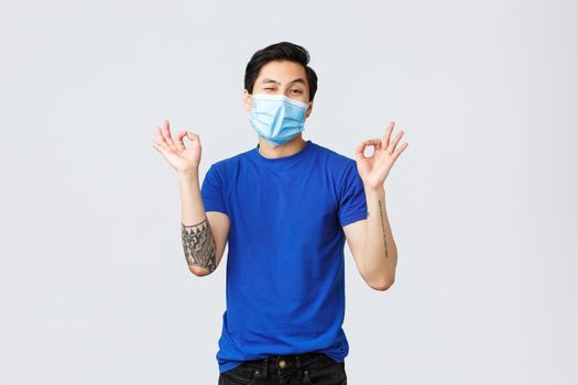 Different emotions, social distancing, self-quarantine on covid-19 and lifestyle concept. Unbothered asian guy acting cool, wear medical mask, show okay, no problem or guarantee sign.
