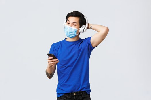 Covid-19 lifestyle, people emotions and leisure on quarantine concept. Handsome young queer asian guy in medical mask and headphones, listening music, using mobile phone.