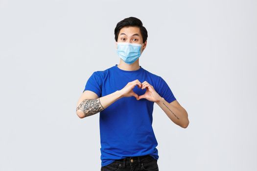 Different emotions, social distancing, self-quarantine on covid-19 and lifestyle concept. Attractive asian man in medical mask showing heart sign over chest to express care or love, sympathy.