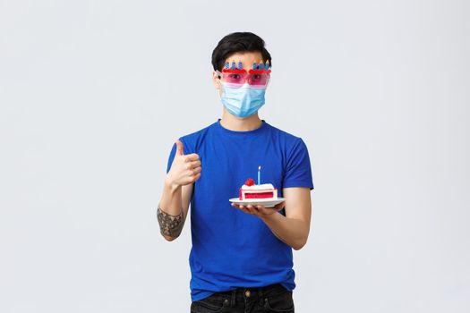 Different emotions, social distancing, self-quarantine on covid-19 and lifestyle concept. Handsome young b-day guy, asian man celebrating birthday in funny glasses, holding cake and thumb-up.