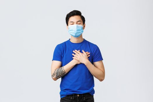Different emotions, social distancing, self-quarantine on coronavirus and lifestyle concept. Dreamy and caring, tender asian man in medical mask, close eyes and touch heart, feel love and warmth.