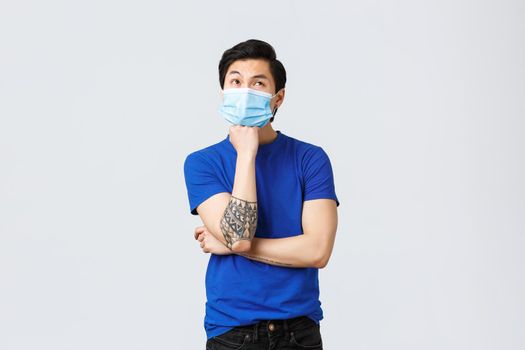 Different emotions, social distancing, self-quarantine on coronavirus and lifestyle concept. Bored and thoughtful asian man in medical mask, lean on arm and look up, thinking, making decision.