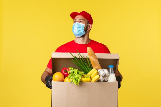 Takeaway delivery, covid-19 quarantine and groceries concept. Carefree handsome courier in red uniform and medical mask, look around with smile, holding box with food order.
