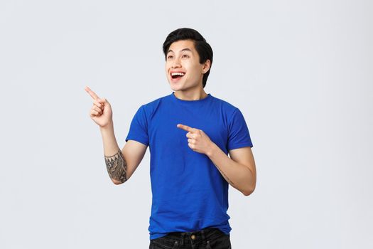 Enthusiastic and happy, good-looking asian guy in blue t-shirt, smiling astonished and amazed, pointing fingers upper left corner, check-out cool advertisement, grey background.