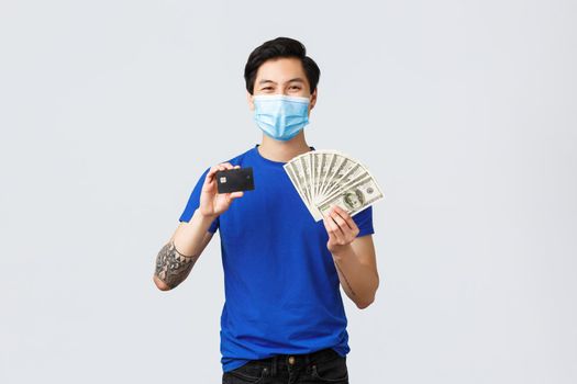 Money, lifestyle, insurance and investment concept. Confident smiling asian man recommend using bank and contactless payment, showing credit card and cash dollars, wear medical mask.