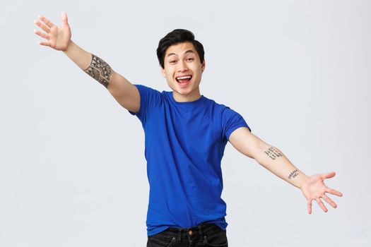 Cheerful handsome asian guy in blue t-shirt greeting friend, spread hands sideways to give cuddles, hugging and welcoming, smiling broadly, standing grey background.