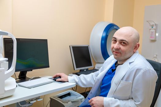 A male ophthalmologist at the workplace at the computer looks at the camera with a smile.
