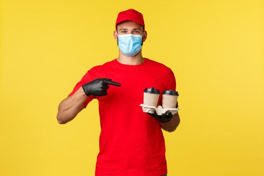 Takeaway delivery, food and drinks, covid-19 quarantine and preventing virus concept. Smiling courier in medical mask and red uniform pointing finger at coffee, beverage order, yellow background.