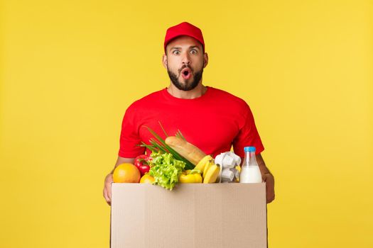 Online delivery, shopping and food order concept. Impressed bearded courier in red uniform cap, t-shirt, say wow amazed as bring grocery box order to customer home, yellow background.