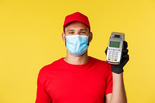 Covid-19, delivery orders, shopping, contactless payment and social distancing concept. Smiling handsome courier in red uniform, medical mask and gloves, giving customer POS terminal.