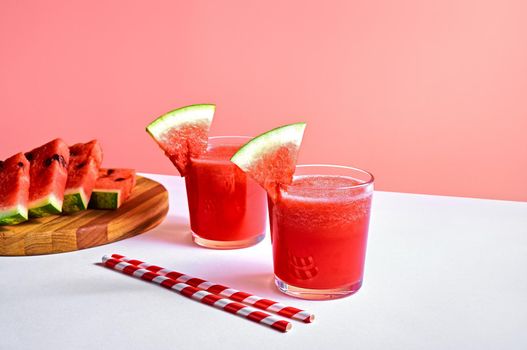 Fresh watermelon juice or smoothie in glasses with watermelon pieces on wooden board on pink background. Refreshing summer drink.