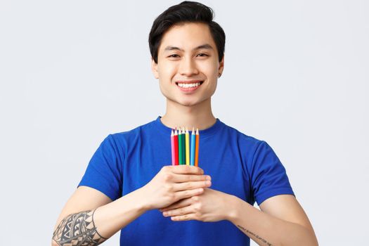 Self-quarantine, social distancine, leisure and lifestyle concept. Young handsome asian man with tattoo show colored pencils, found new hobby feeling happy, smiling at camera.