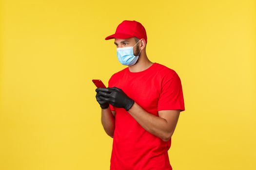 Food delivery, tracking orders, covid-19 and self-quarantine concept. Courier or employee in red uniform, medical mask and gloves, using smartphone, looking left, search client adress to deliver.