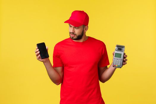 Contactless payment, online delivery, internet shopping concept. Curious courier in red uniform cap and t-shirt, looking at mobile phone, holding smartphone and POS terminal, yellow background.