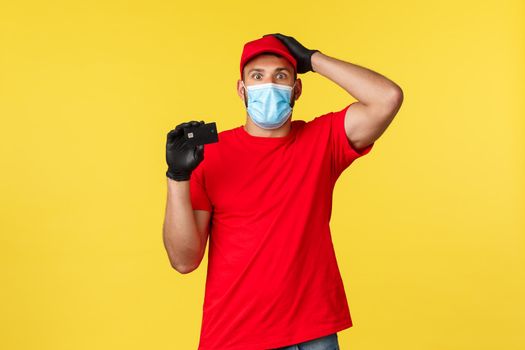 Express delivery during pandemic, covid-19, safe shipping, online shopping concept. Confused and clueless courier in medical mask, gloves and uniform, scratch head unsure, showing credit card.