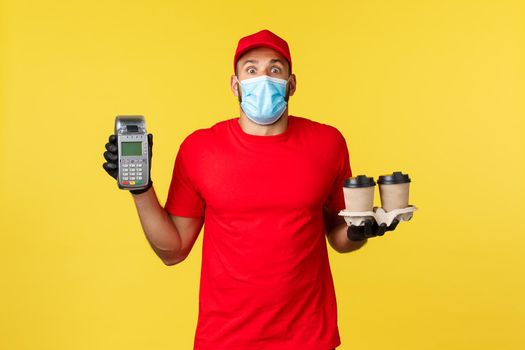Takeaway delivery, drinks, covid-19 quarantine and preventing virus concept. Surprised gasping courier in red uniform and medical mask, startled stare camera, show POS terminal and coffee order.