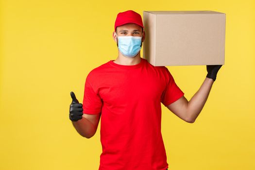 Express delivery during pandemic, covid-19, safe shipping, online shopping concept. Strong young courier in red uniform of company, hold box on shoulder and thumb-up, guarantee safety of parcel.