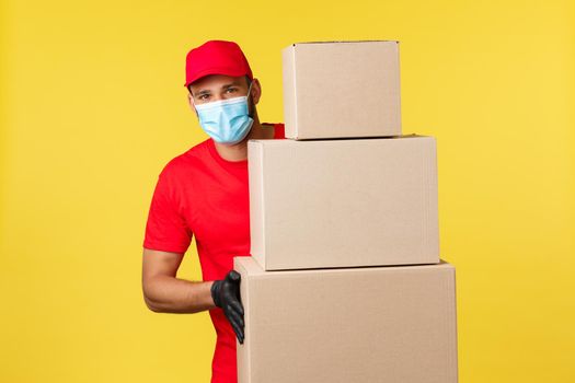 Express delivery during pandemic, covid-19, safe shipping, online shopping concept. Enthusiastic young courier in red uniform, medical mask, standing near piles of boxes with client orders.