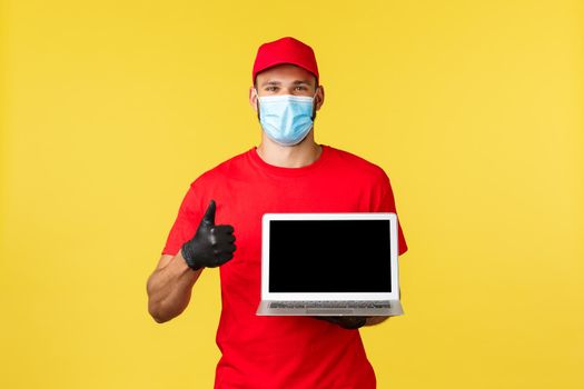 covid-19, safe shopping, express delivery and order tracking concept. Friendly young courier in red uniform and medical mask, show thumb-up, recommend visit link or site, show laptop screen.