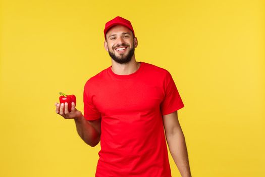 Online delivery, shopping and food order concept. Handsome cheerful courier in red uniform cap and t-shirt, holding fresh pepper, deliver groceries from shop to customer home.