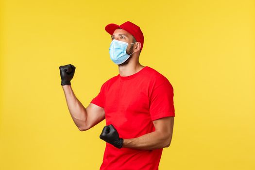 Express delivery during pandemic, covid-19, safe shipping, online shopping concept. Rejoicing happy courier in red uniform, medical mask and gloves, fist pump look upper left at screen, celebrating.