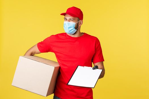 Express delivery during pandemic, covid-19, safe shipping, shopping concept. Enthusiastic handsome courier in red uniform, medical mask and gloves, hold package box and give clipboard form to sign.
