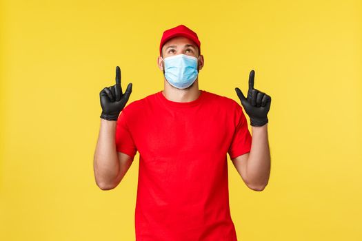 Express delivery during pandemic, covid-19, safe shipping, shopping concept. Satisfied smiling courier in red uniform, medical mask and gloves, pointing and looking up at promo, yellow background.