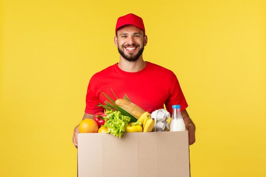 Online delivery, shopping and food order concept. Nice delivery guy, courier in red uniform company, bring grocery order in box to client, smilign friendly, standing yellow background.