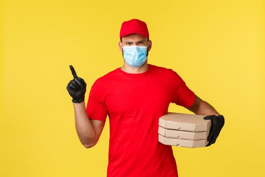 Food delivery, tracking orders, covid-19 and self-quarantine concept. Displeased serious courier in red uniform, medical mask and gloves frowning bothered, pointing left, holding pizza order.