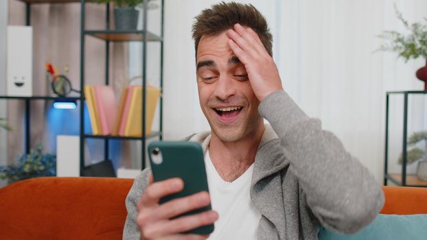 Excited happy caucasian man use mobile cell phone typing browsing say Wow yes found out great big win good news celebrate doing winner gesture. Young adult guy at home in living room sitting on sofa