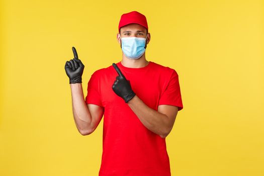 Express delivery during pandemic, covid-19, safe shipping, online shopping concept. Smiling courier in medical mask and red uniform cap, t-shirt, pointing fingers upper left corner, show banner.