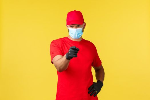 Express delivery during pandemic, covid-19, safe shipping, online shopping concept. Handsome courier in red uniform, medical mask and gloves, pointing client and make phone call sign.