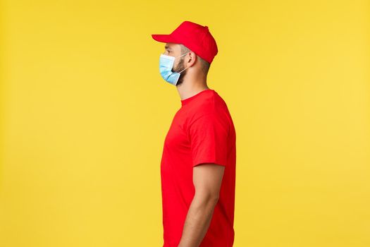 Express delivery during pandemic, covid-19, safe shipping, online shopping concept. Profile of cheerful courier in red uniform and medical mask looking left, work with clients, taking package.