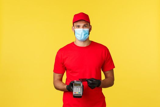 Food delivery, tracking orders, covid-19 and self-quarantine concept. Friendly courier in red uniform, medical mask and gloves, using credit card on POS terminal to pay for order contactless.