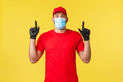 Express delivery during pandemic, covid-19, safe shipping, online shopping concept. Impressed and excited courier in red uniform, medical mask and gloves pointing up, look astonished.
