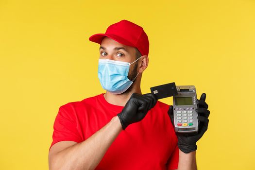 Covid-19, delivery orders, shopping, contactless payment and social distancing concept. Cheerful courier in red uniform and medical mask, gloves press credit card to payment terminal.