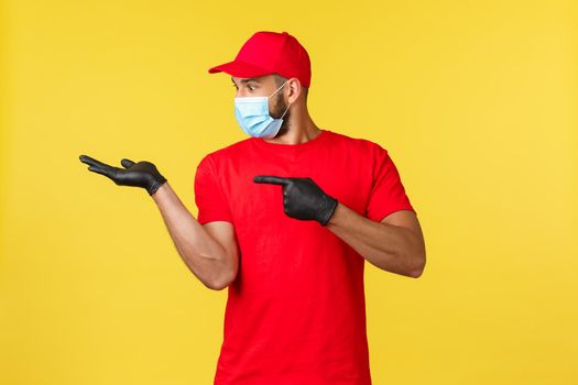 Express delivery during pandemic, covid-19, safe shipping, online shopping concept. Excited happy courier in red uniform and medical mask, gloves, pointing finger and looking at palm with product.