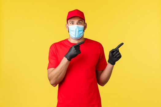 Express delivery during pandemic, covid-19, safe shipping, online shopping concept. Excited courier in red uniform, merical mask and gloves, pointing right, tell big news.