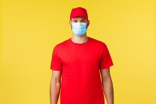 Express delivery during pandemic, covid-19, safe shipping, online shopping concept. Young courier in red uniform standing over yellow background, deliver orders during quarantine pandemic times.
