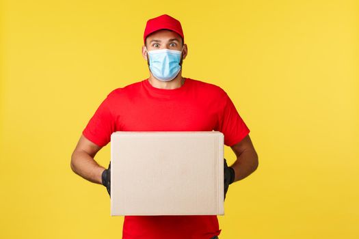 Express delivery during pandemic, covid-19, safe shipping, online shopping concept. Enthusiastic young courier in red uniform and medical mask, look astonished, holding client order box.