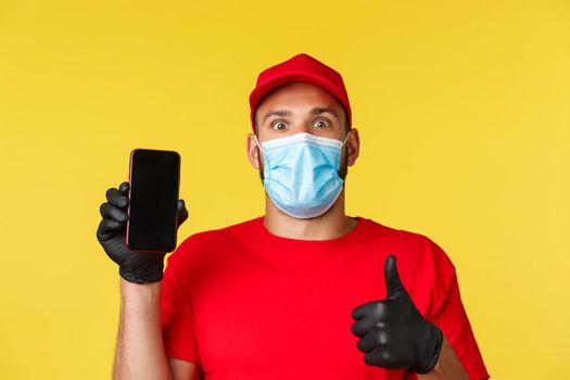 Covid-19, delivery orders, shopping, contactless payment and social distancing concept. Excited employee, courier in red uniform and medical mask, impressed with new app, show mobile phone screen.