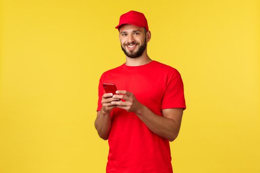 Contactless payment, online delivery, internet shopping and parcel tracking concept. Friendly smiling courier in red uniform cap, t-shirt, using mobile phone and look camera, yellow background.