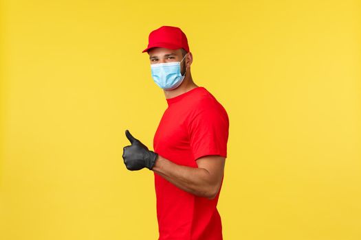 Express delivery during pandemic, covid-19, safe shipping, online shopping concept. Friendly male courier in red uniform and medical mask, provide safe contactless package deliver, thumb-up.