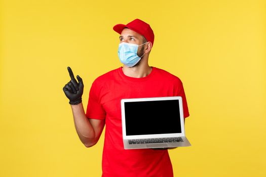covid-19, safe shopping, express delivery and order tracking concept. Young courier in red uniform of transportation service, showing laptop screen and pointing upper left corner.